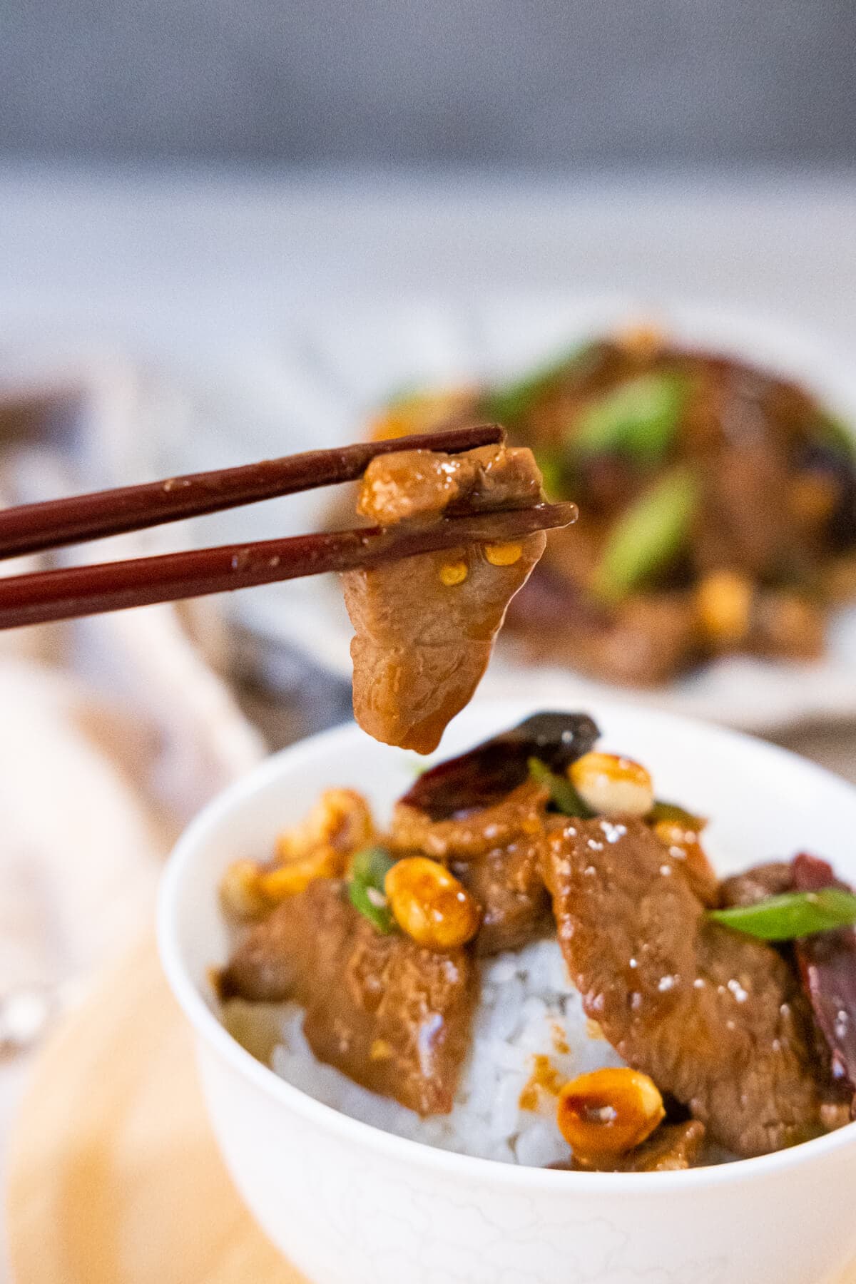 A chopstick holding up a slice of beed tenderloin coated with Kung Pao sauce from a bowl of rice topped with Kung Pao Beef in a bowl. 