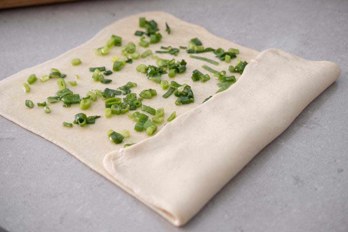 A flat, rectangle bun dough is folded from one side to the center and has chopped scallions evenly spread on it.