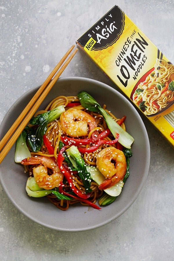 Lo mein in a bowl covered in shrimp and vegetables, next to a box of the lo mein noodles from McCormick Simply Asia.