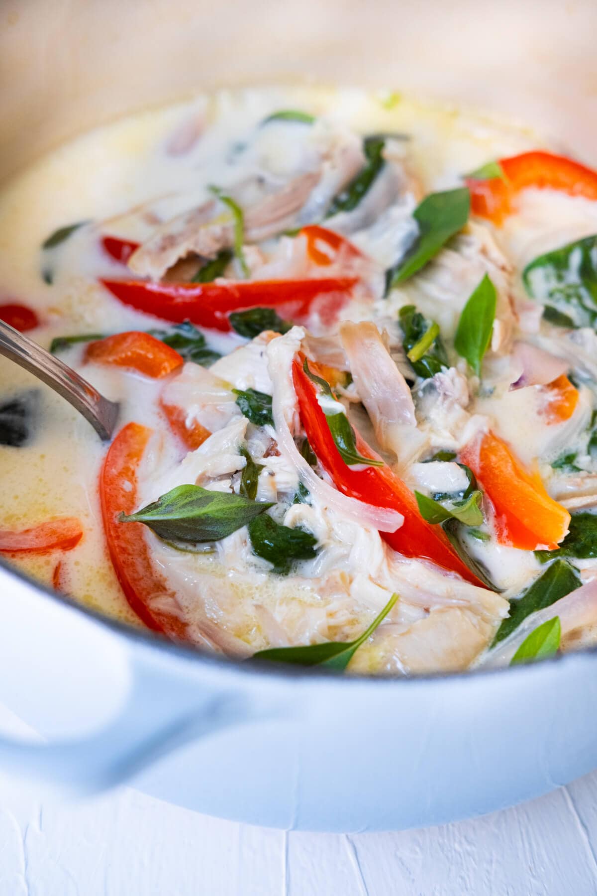 Stew in a pot with coconut milk, chicken, red bell pepper, baby spinach leaves and basil leaves on top.
